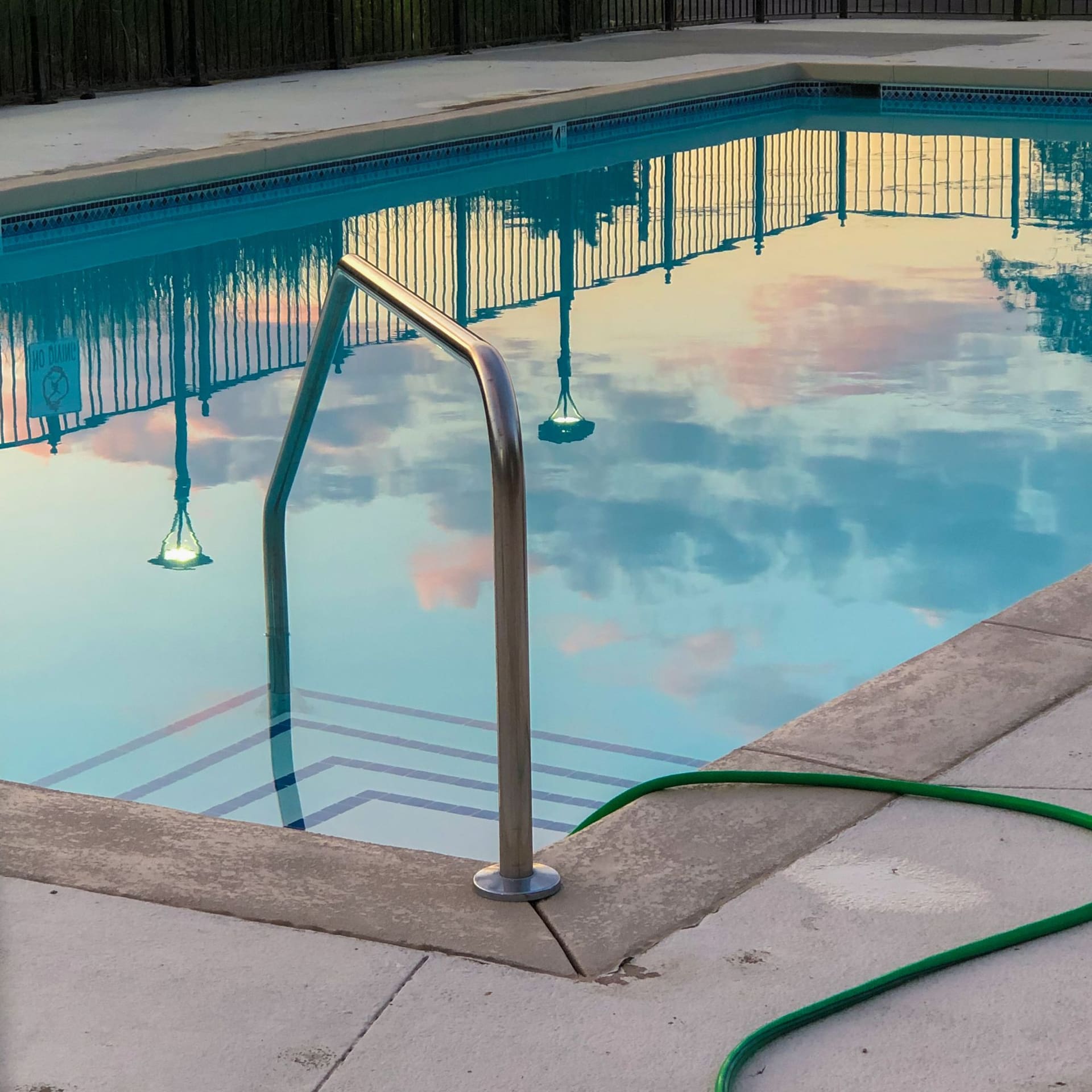 Calm pool water reflecting the sky, with a steel ladder and a garden hose on the deck
